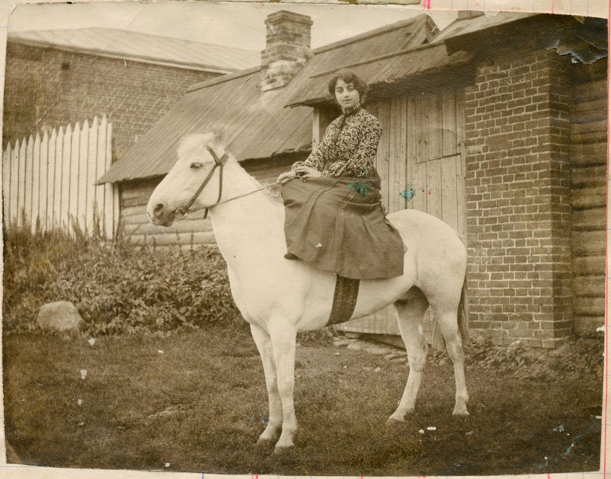 Motivet viser en ung kvinne som rir på en hvit hest. Omgivelsene er landlige og med en synlig tømmerbygning, skorstein og hvitt stakittgjerde i bakgrunnen.