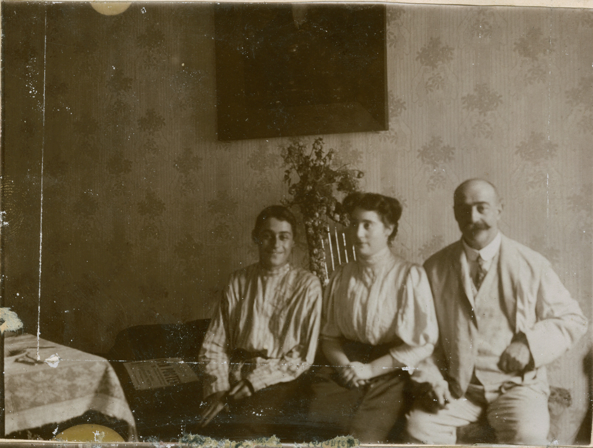 Søsknene Valentine og Ignazy avbildet sammen med sin far, Mikhail Rostin. De sitter på en divan med en blomsterdekorert harpe i bakgrunnen. På den tapetserte veggen bak henger et maleri.