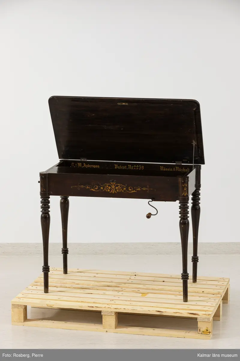 KLM 25434. Pianoharpa, vevpiano. Rektangulärt brunbetsat och lackerat bord med svarvade ben. På sargen schablonmåleri i guldfärg. Bordsskivan kan öppas, inuti en inbyggd melodivals som drivs med en löstagbar vev. Valsen kan justeras i sidled för att spela olika melodier. Inuti text i guld: G. & W. Andersson Patent No 2239 Nässja & Näshult. Intill valsen en papperslapp med text: No 89.