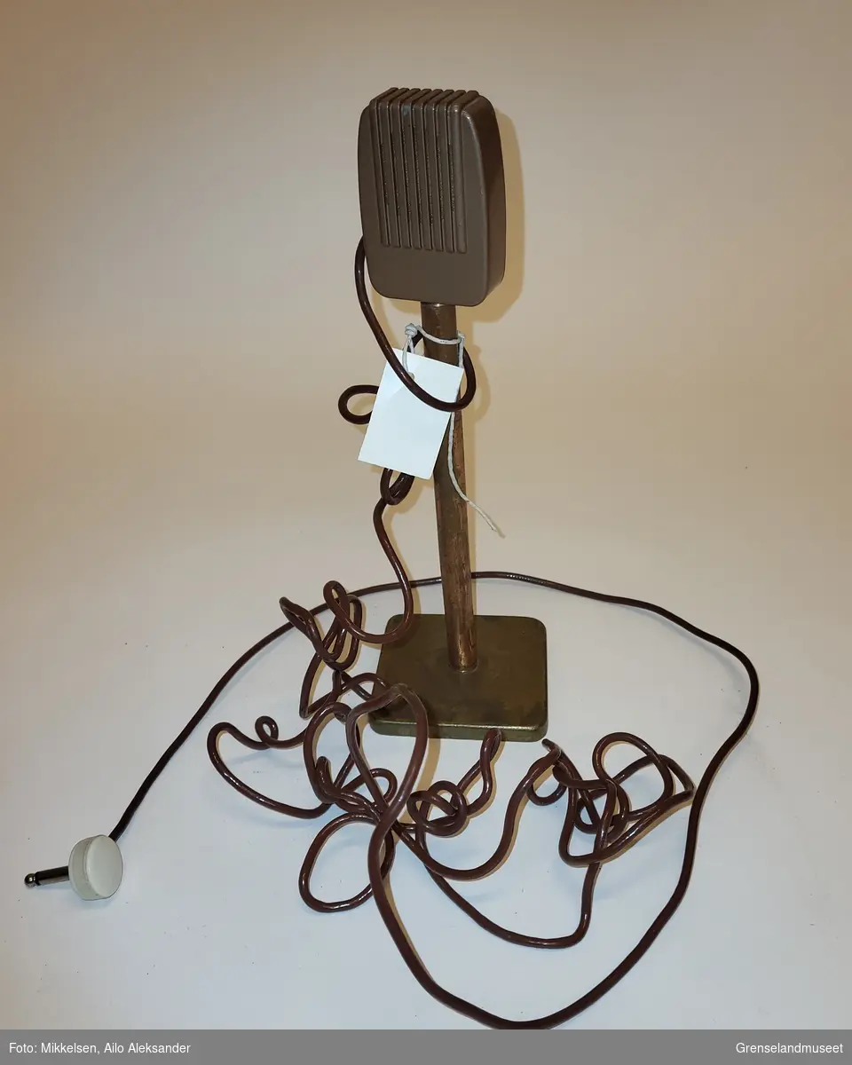 Gjenstanden er en mikrofon stående på en firkantet messing fot. Messingen har begynt å oksidere og viser tegn på alderstidsbruk. Mikrofon ledningen er brun og svært lang med et lydsstøpsel.  