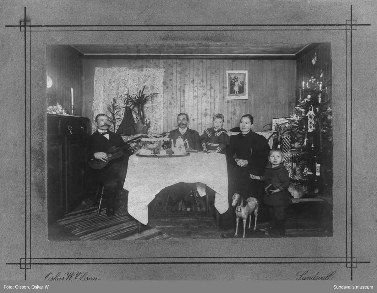 Två män, en kvinna och två barn vid ett bord med dukad kaffebricka. En av männen knäpper på en gitarr. En pyntad julgran står vid ena sidan. Ett av barnen har en stor leksakshäst i trä. På baksidan står: Familj i Torpshammar. Ur Anders Nylanders samling.