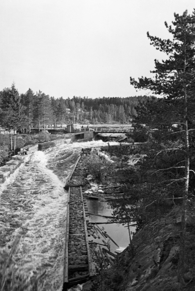 Haldammen ved Ytre Halåas utløp fra Halsjøen lengst øst i Våler kommune i Hedmark. Fotografiet er tatt i motstrøms retning, fra østre elvebredd i begynnelsen av juni 1940. Dammen sto åpen, så vannet strømmet ut i et elveløp som var omgitt av skådammer - en natursteinsmur på vestsida og en steinfylt tømmerkistekonstruksjon på østsida. Her foregikk det åpenbart tømmerfløting.