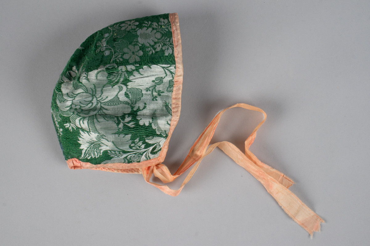 Liten håndsydd dåpslue i grønn silkebrokade og rosa kantbånd og knytebånd i silke. Lua er satt sammen av seks deler og har hvit foring av glanset linlerret