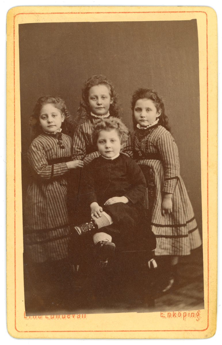 Svartvit fotografi av fyra barn. En pojke sittande på en stol framför tre stående flickor i liknande kläder.