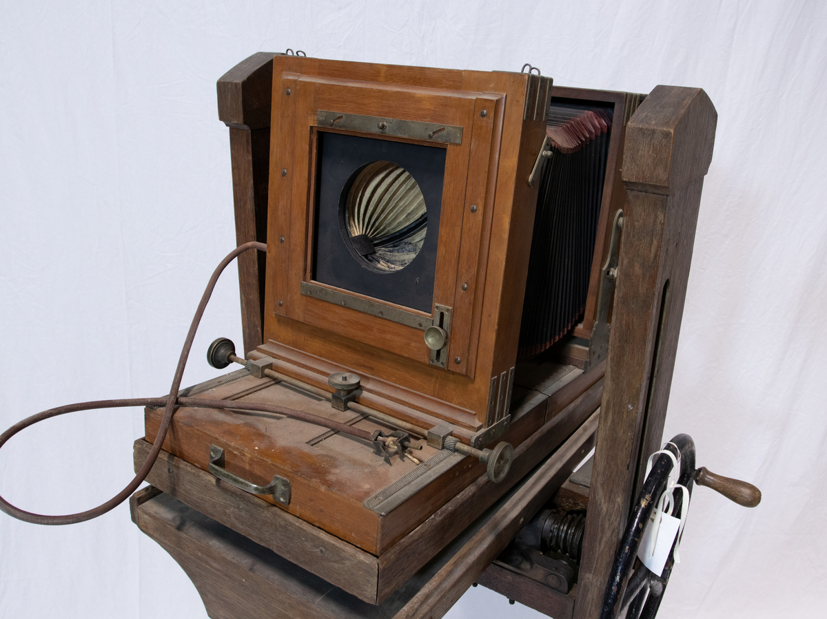 Fotografiapparat på stativ med  gjengeverk og drivhjul drevet av en sveis. Stativet har en vippeplate forregulering av fotokasse.