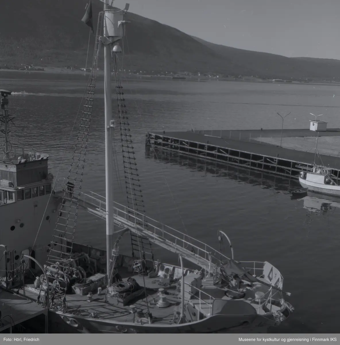 Bildet viser baugen til en fangstbåt, gjenkjennelig ved harpunkanonen og utsiktstønna. Båten ligger i Tromsø havn. Tromsøysundet og fjell ligger i bakgrunnen.