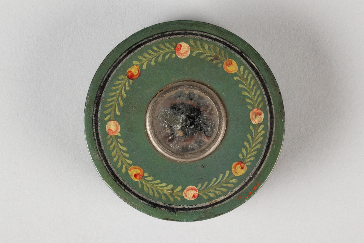 Lav, sylinderformet, grønnmalt pudderdåse i tre med et lite sirkulært speil på lokket. Lokket er dekorert med plantemotiv
