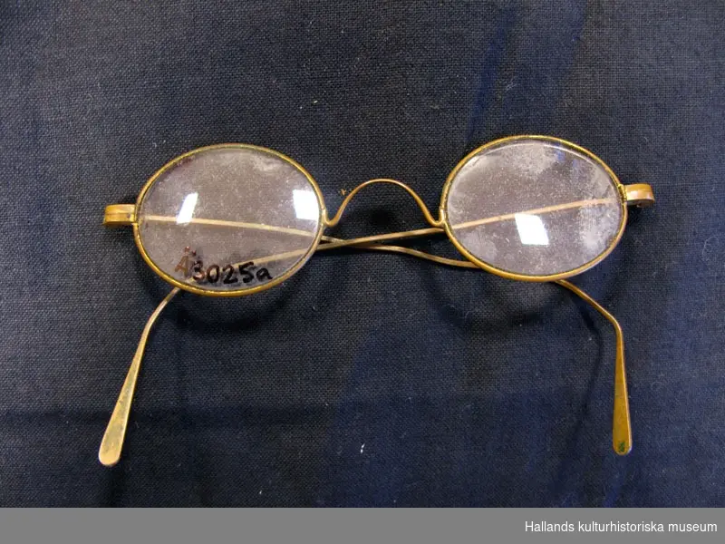 Glasögon med fodral. Glasögon med ovala glas och bågar i någon gulmetall, kanske mässing. Lite böjda, lite erjade. Fodral i papp, oval form, svart och brunt i färgen.