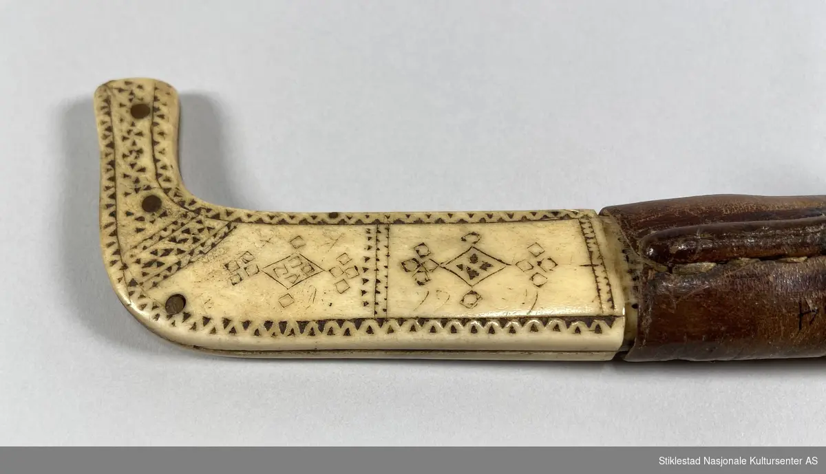 Knivslire laget av reinshorn og lær. Klinket med kobbertråd. Messingmaljer øverst på slirelæret som dekor. Inspirert av samisk dekor. Knivsliret er merket med I.J. 1934
