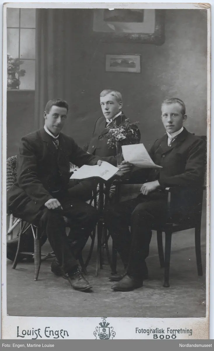 Portrett av tre unge menn. Atelierbilde. De sitter rund et bord med dokumenter i hånden. Kan være sønner til kjøpmann Ludvig Fremmerlid på Bogøy handelssted. Bakerst skal være Nils Fremmerlid, f. 1892.