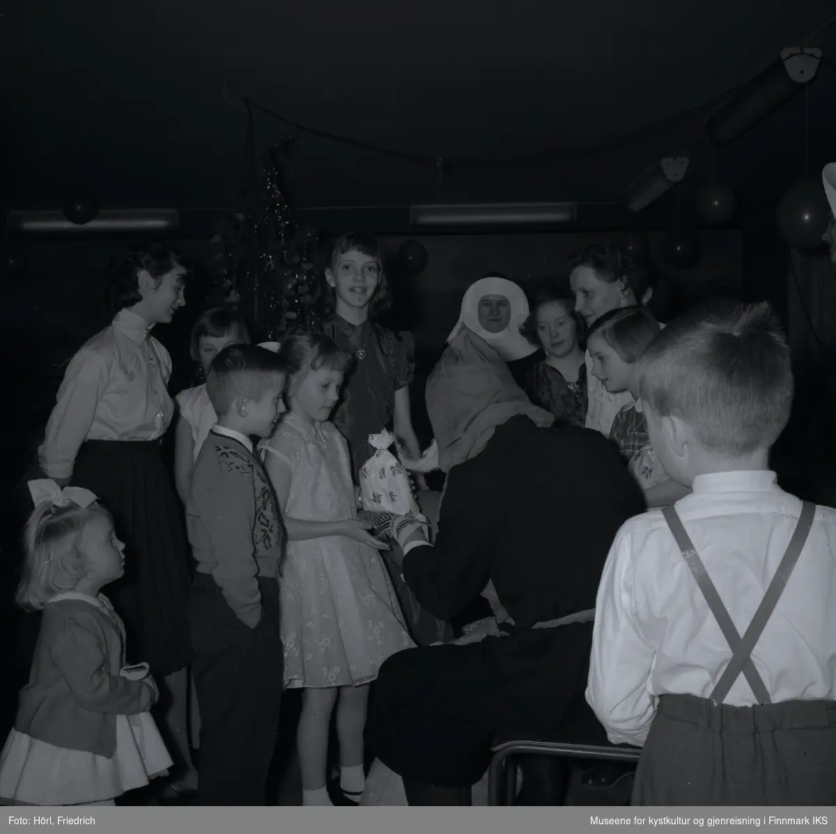 Barn og kvinner omringer julenissen på juletrefesten i den katolske menigheten i Hammerfest i 1957. Julenissen overrekker en gave til en jente. Hun er festlig kledd i en fin kjole og pensko. Ved siden av henne står det en gutt som er svært opprømt. Bak henne ser man en eldre jente som smiler glad til fotografen. Jenta nederst til venstre i bildet er Wenche Rose Trondsen. I bakgrunnen ser man juletreet. Girlander med ballonger henger fra taket.