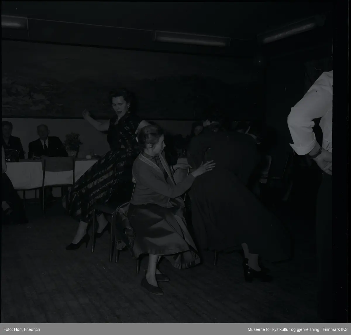 Den katolske menigheten i Hammerfest feirer juletrefest i 1957. Det er damerunde i stolleken og engasjerte deltagere prøver å finne seg en plass når musikken slutter. Andre sitter ved bordene og ser på. På veggen i bakgrunnen er det et stort landskapsmaleri.