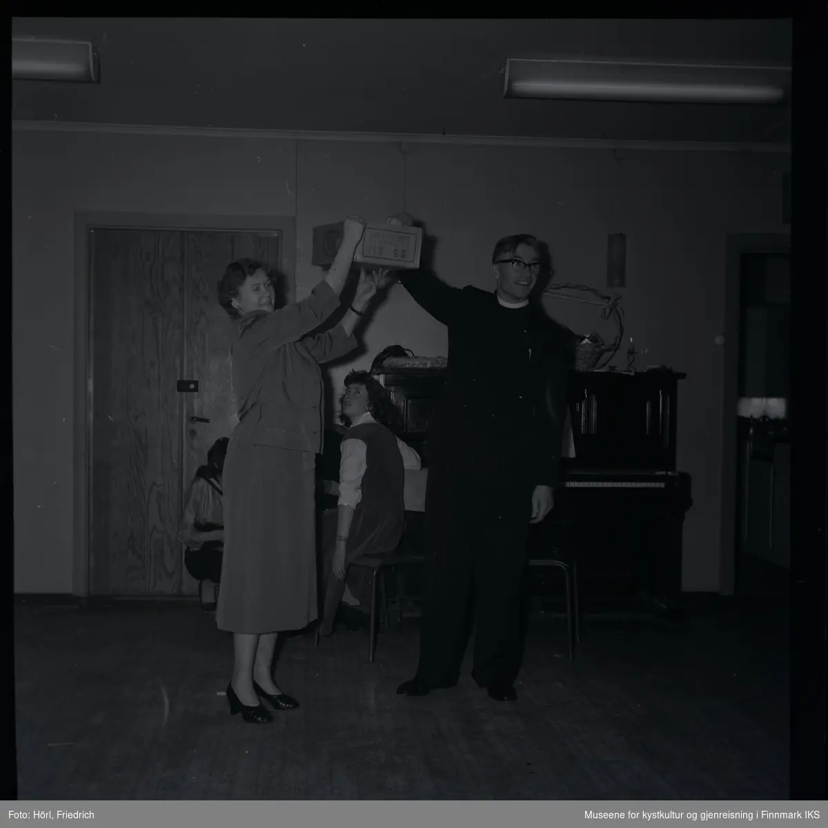 Den katolske menigheten i Hammerfest feirer juletrefest i 1957. Bildet viser en kvinne som holder opp en eske. Pater Spitz stikker hånden inn i den for å trekke en lodd. I bakgrunnen sitter tre kvinner ved siden av et piano, en av dem ser opp til esken med loddene. På pianoet ligger noe som ser ut som et tysk julebrød kalt "Stollen", der står det også en kurv med sløyfe og et stearinlys.