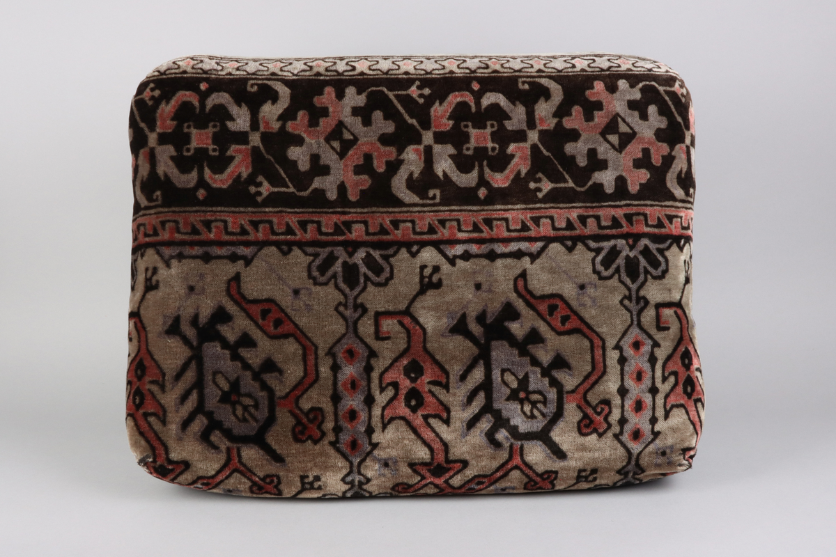 En kudde av mönstrad schagg. Orientaliserande mönster i färgerna grå, svart och vinröd. Del i en grupp med överkast och kuddar till en sk. Ottoman, en typ av bäddsoffa.
