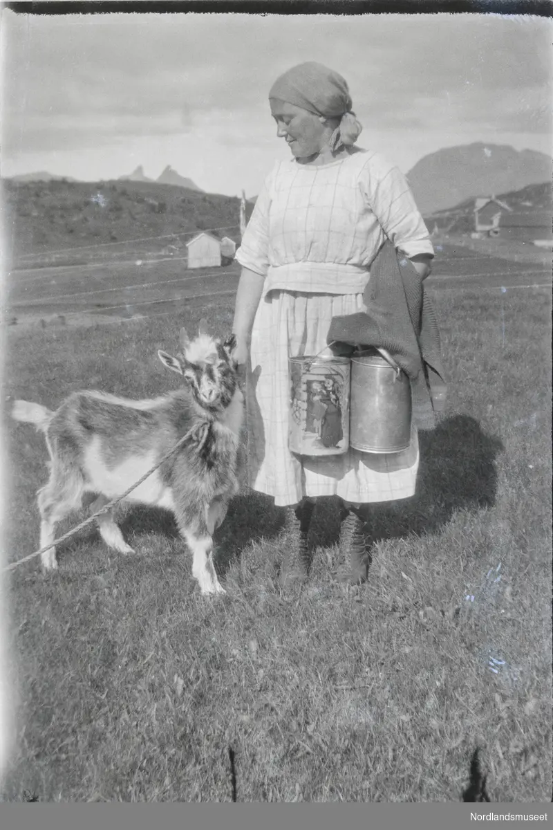 En kvinne med skaut i lys rutet kjole og to melkespann i handa og en geit.