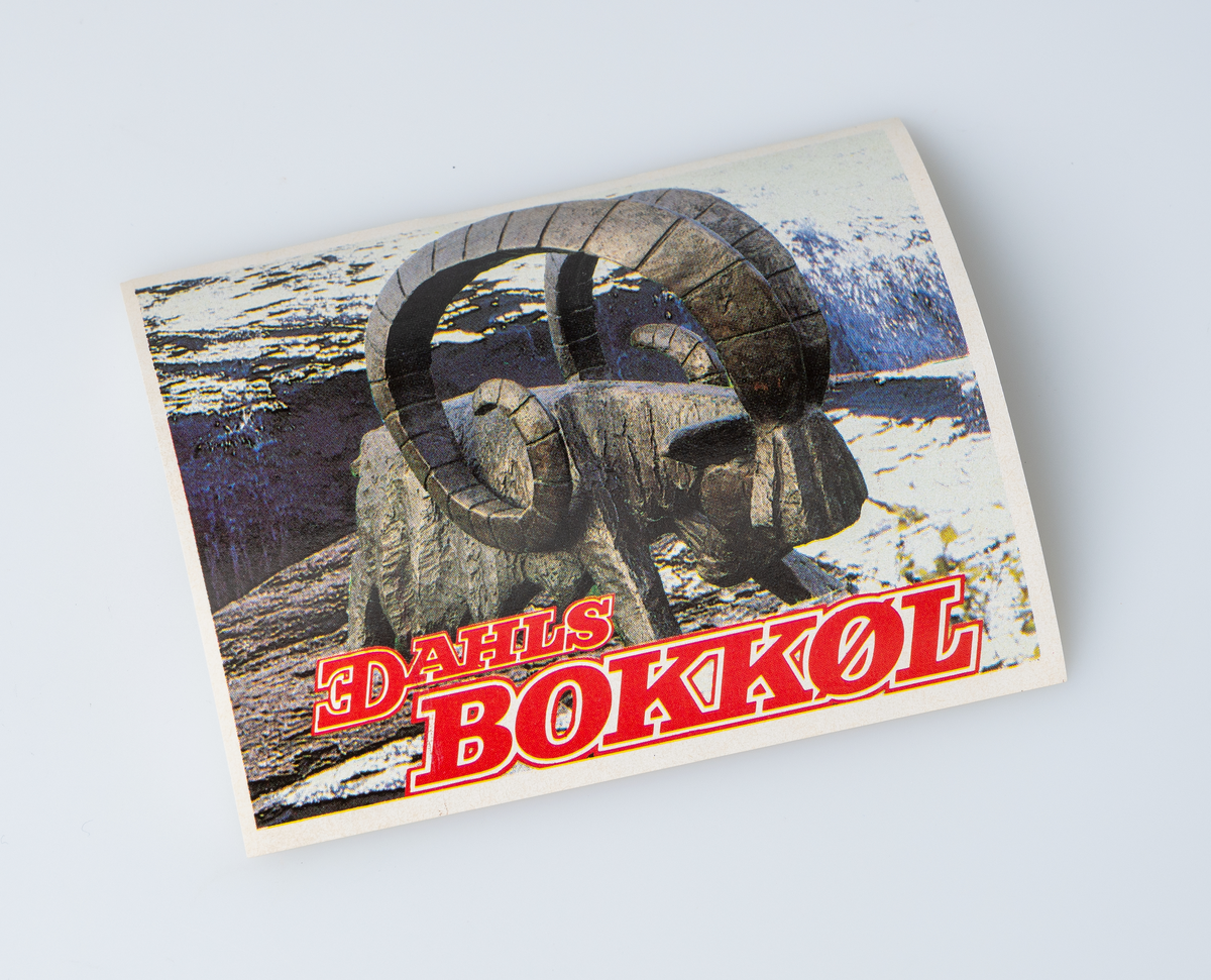 En firkantet øletikett av papir. Etiketten er til E. C. Dahls Bokkøl. Motivet på etiketten er et fotografi av en statue av en bukk som står i et delvis snødekt fjellandskap. Nederst er det tekst med røde bokstaver.