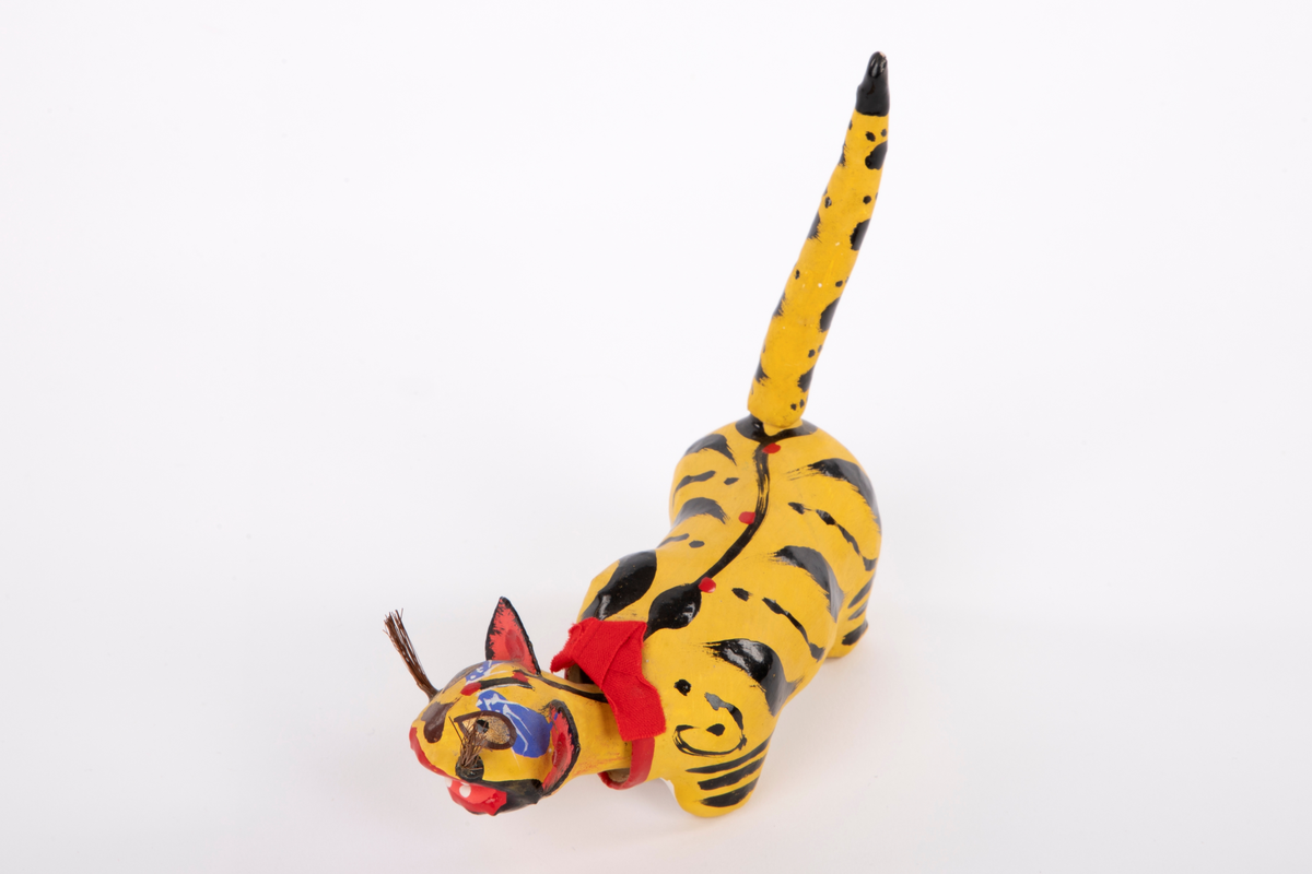 Tiger i pappmasjé med bevegelig hode. Tigeren er gul med svarte striper, har lang kropp med løs hale og et rødt bånd rundt nakken. Figuren har utstående ører, værhår og åpen munn med hvite tenner.