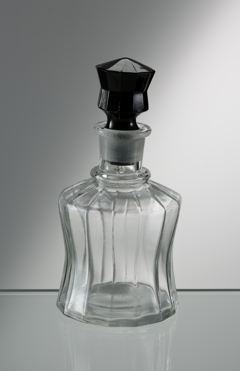 Oljeflaska tillhörande ett bordskryddställ (M 68314-1 till M 68314-6) på fat i sammanlagt sex delar (fatet inkluderat). Timglasformad flaska med stående ribbor. Svart propp, även den timglasformad med stående ribbor.