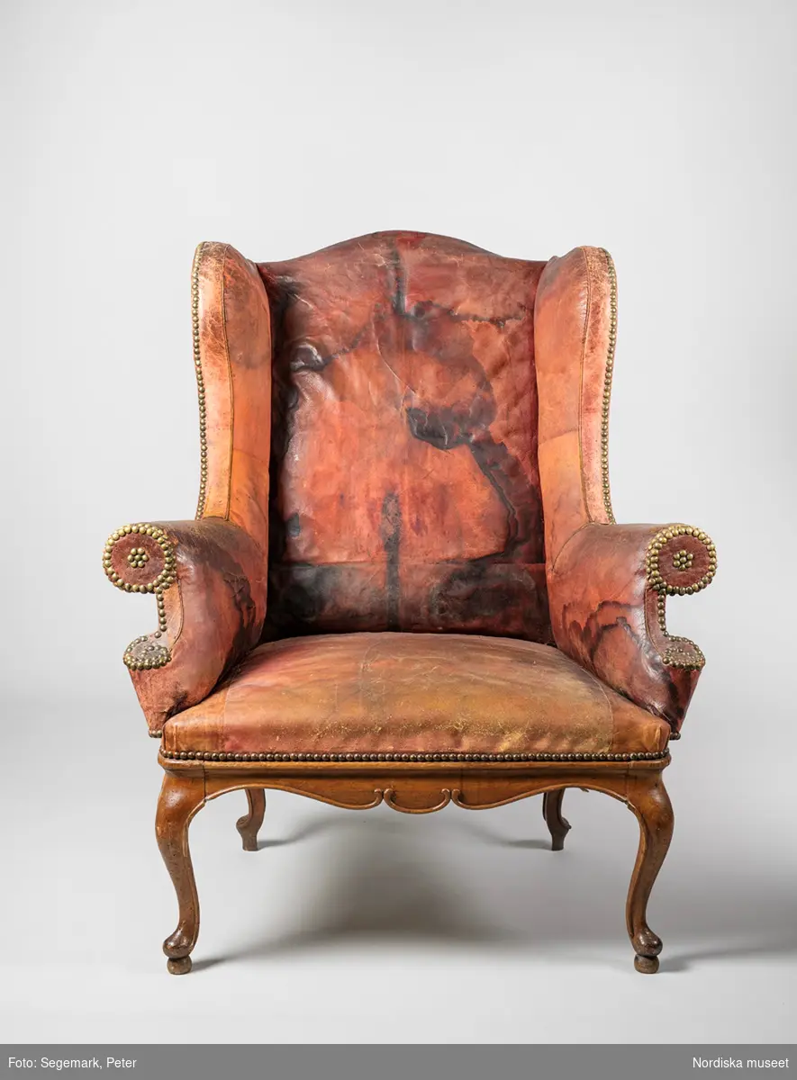 Fåtölj med "öronlappar", även kallad sovstol, rokoko, omkring 1750, sannolikt tillverkad i Stockholm efter engelsk modell. Helt klädd med rött skinn (marokäng/saffian), fläckat och kraftigt blekt. Skinnklädseln fästad med tätt sittande möbelspik av mässing. Underrede av brunbonad (ursprungligen rödfärgad) al. Ryggens insida har stora, mörka fläckar. 
Anm: Renoverad. Sitsen moderniserad med resårstoppning. Bakbenen utbytta mot ben av ek. Sliten skinnklädsel.
/Anna Arfvidsson Womack 2021