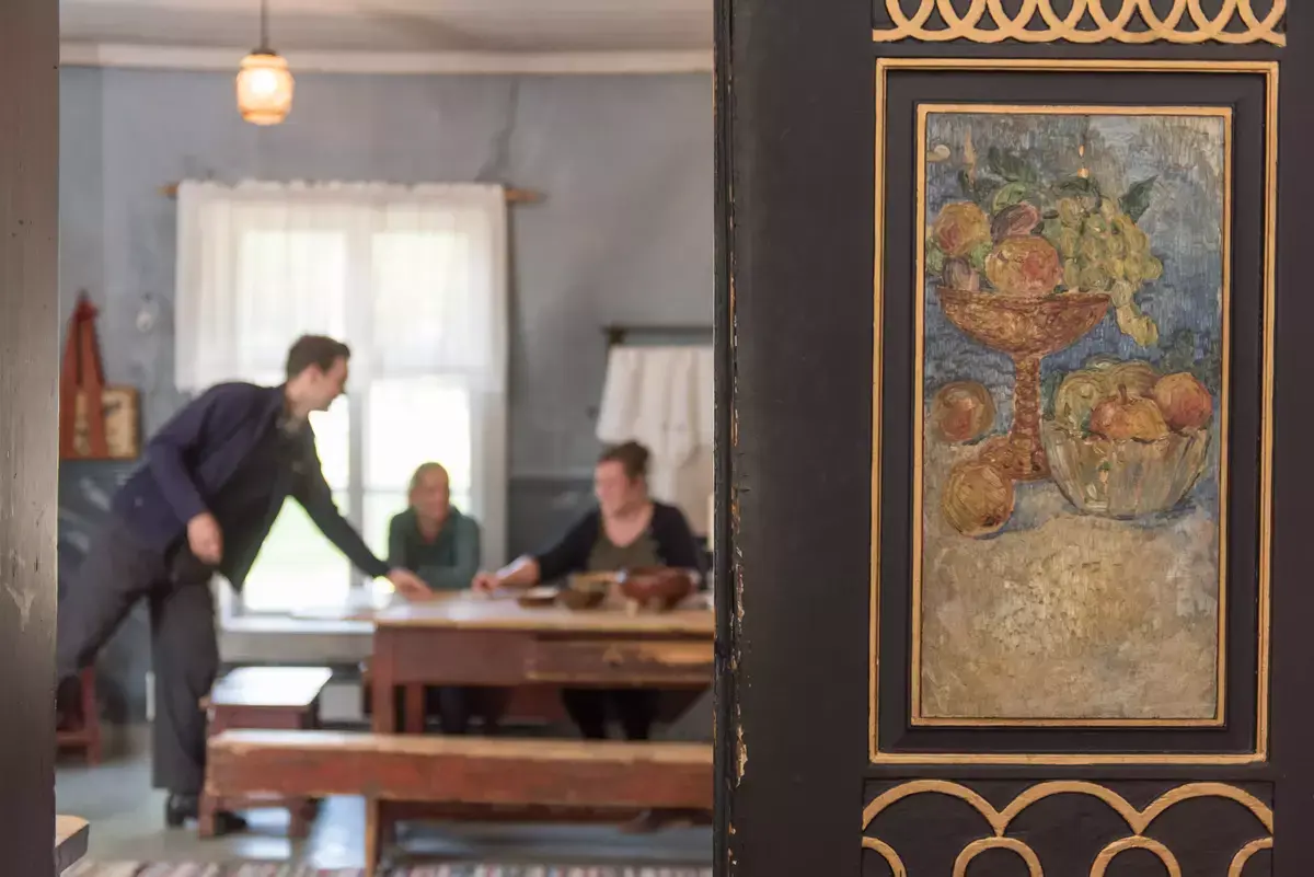 markedsføringsfoto tatt gjennom dør inn på det gamle kjøkkenet på Gjøvik gård. Gammelt langbord og langkrakker av tre. To personer sitter ved bordet og en tredje person skjenker kaffe.