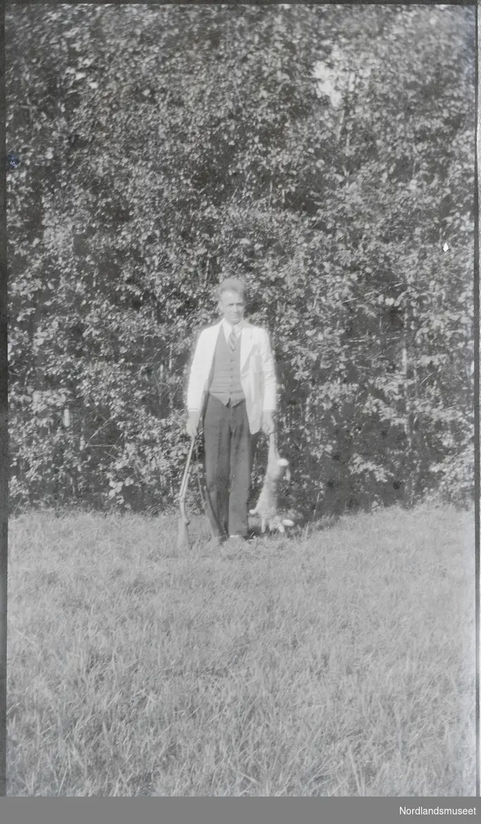Mann i bukse, veste, slips og hvit dressjakke, frakk, kittel, med en død hare i den ene hånda og ei hagle i den andre.