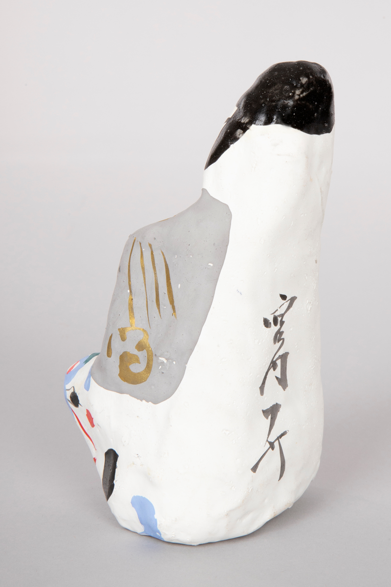 Figur i pappmasjé som forestiller en stor rytter på en liten okse. Mannen har på seg rød og grå kappe med gullbånd. Japanske tegn på baksiden av figuren.