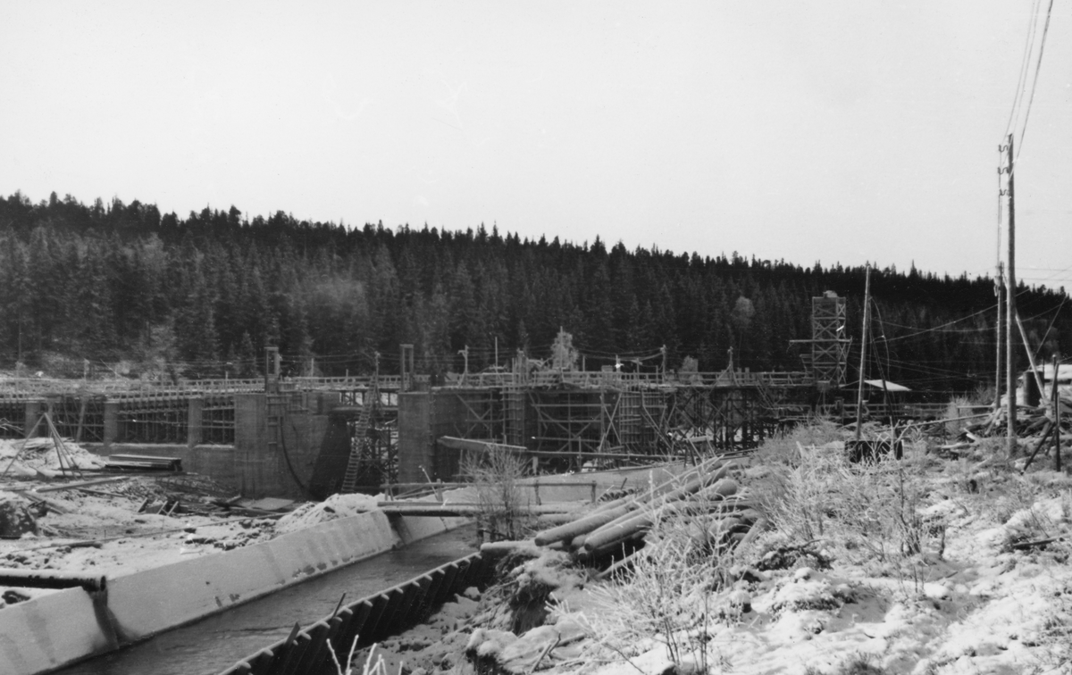 Bygging av reguleringsdam ved elva Søndre Osas utløp av Osensjøen i Åmot i Hedmark seinhøstes i 1937. Dette ble gjort for å kunne bruke sjøen som vannreservoar for elvekraftverk i den nedenforliggende delen av Glommavassdraget ved å tappe vann i perioder da det naturlige tilsiget var beskjedent, som for eksempel i vintersesongen. Dette fotografiet er tatt fra søndre elvebredd, på nedstrøms side av damstedet, på et tidspunkt da den nedre delen av dammen var langt på veg ferdigstøpt. I forgrunnen ser vi ei renne på elvebrinken, der vannet fra sjøen inntil videre kunne renne uten at det forstyrret anleggsarbeidet.
