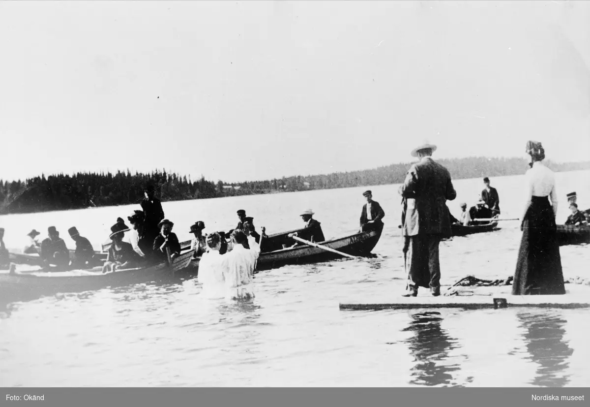 En grupp människor är samlade i båtar och på en brygga. I vattnet står två vitklädda personer. Bilden visare det första baptistdopet i Tåsjötrakten 1918. Baptismen bredde ut sig i Ångermanland från slutet av 1800-talet.
