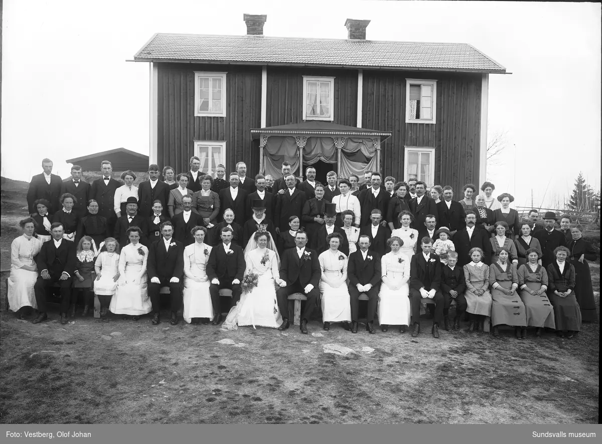 Gruppbild från ett bröllop med brudpar och bröllopsgäster framför en lantligt belägen rödmålad bostadsbyggnad i två våningar och med tyg draperat på brokvisten.