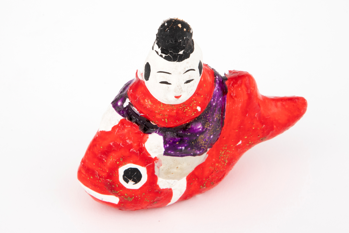 Figur i pappmasjé og gips som forestiller en mann ridende på en fisk. Fisken er rød og blank, mens mannen er kledd i en drakt i grå, lilla og rød med gullglitter. Hvit bakside med japanske tegn.