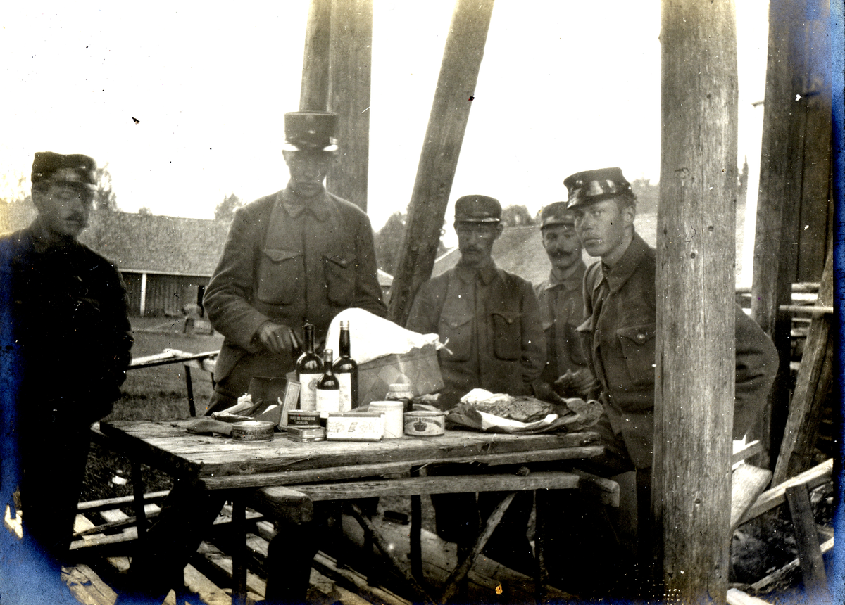 Soldater i sitter ved et bord med mat og drikke på. 1905.
Da Lindallskurv var kommet.
Bilde er fra fotoalbum GM.036887.
