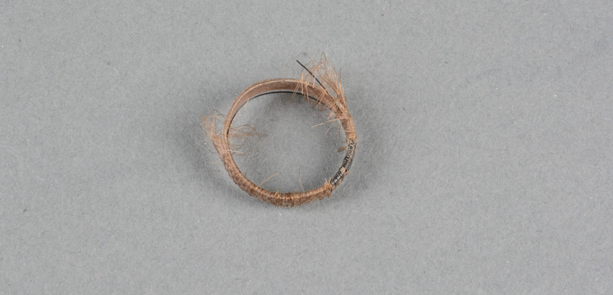 Fingerring av hår og horn, flettet rundt flat, stiv hornring. Innslag av sort hår i mønster. På halve ringen er hårflettingen slitt av.