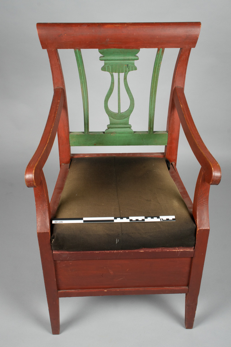 Nattstol eller dostol i tre med armlener og utskåret rygg. Avtagbart polstret sete trukket i tøy. Lite rundt lokk med tøyhåndtak under setet, over åpning.