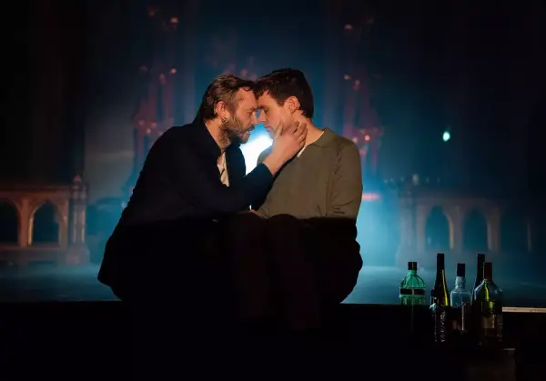 To menn sitter i mørke oppå et flygel, mannen til venstre tar forsiktig og kjærlig på ansiktet til mannen til høyre, de opplyses av forsiktig lys bakfra.