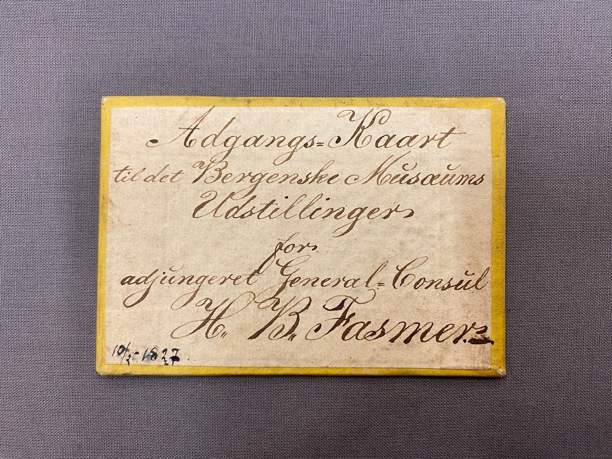 Adgangskort til de første utstillinger ved Bergen Museum. Håndskrevet tekst på den ene siden, ensfarget gul flate på den andre siden med lakkstempel midt på.