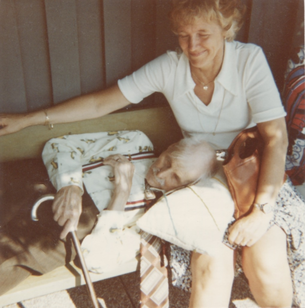 Karl-Erik "Masen" Andersson ligger på en träbänk och vilar huvudet i knäet på en kvinna, Brattåshemmet 1970-tal. I handen håller Karl-Erik en käpp.