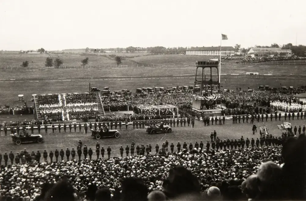 Fra tribunen fylt med tilskuere ser vi mot en parade av biler med æresvakter på begge sider av traséen. Tilskuerne som sitter på tribunen på motsatt side danner et stort norsk flagg. Bak dem står flere tilskuere eller deltakere og en rekke biler. Et tårn er reist til høyre i bildet og har påmontert en flaggstang med amerikansk flagg. I bakgrunnen til høyre to lange, brakkelignende bygninger.