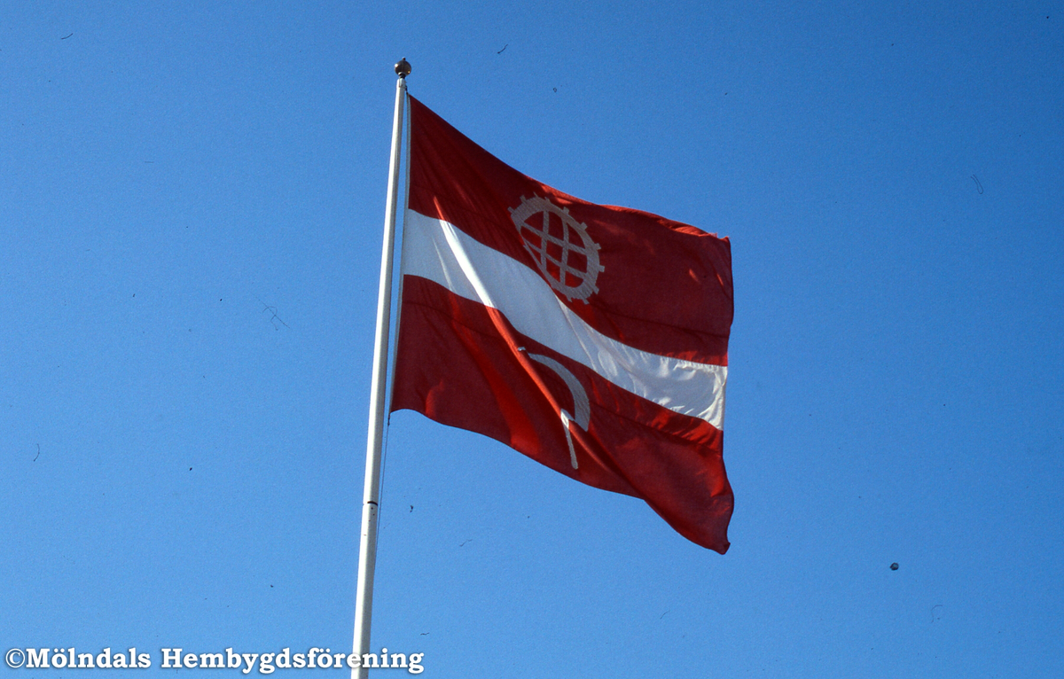 Den första kommunflaggan invigdes 24/6 1976. En kommunfullmäktigeordförande, Åke Baaz, tyckte den var i rödaste laget och beställde en ny. Den gamla brändes upp. Fotografi av Mölndals kommunflagga vid stadshuset i Mölndal.