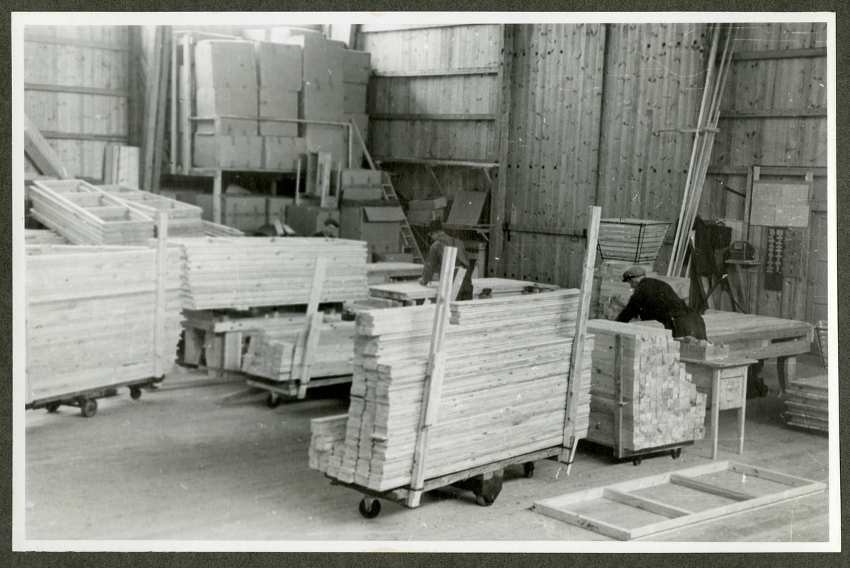 Foto av tremateriale på paller og og menn ved snekkerbenker på  Aall-Ulefoss Brugs Dørfabrikk