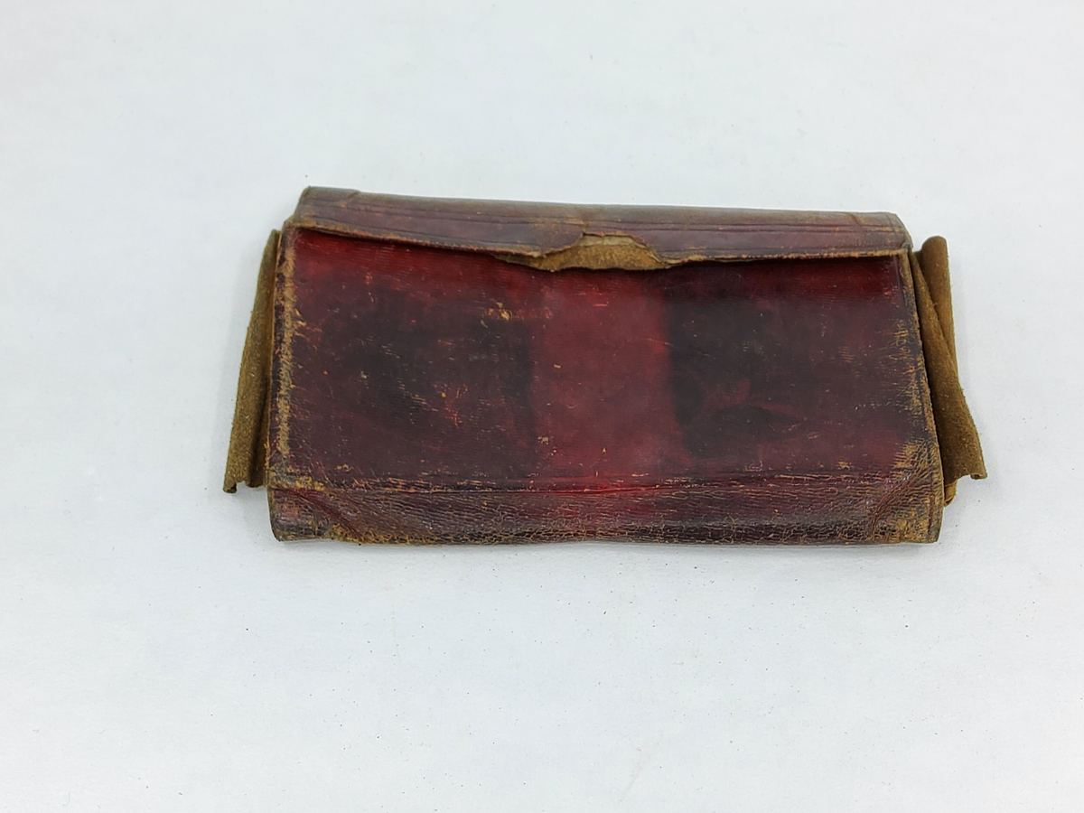 Plånbok av röd saffian, två fack av papp med tryckta mönster på framsidan. Foder av tunt skinn på mittdelen och i sidorna. Illa medfaren, rem saknas.