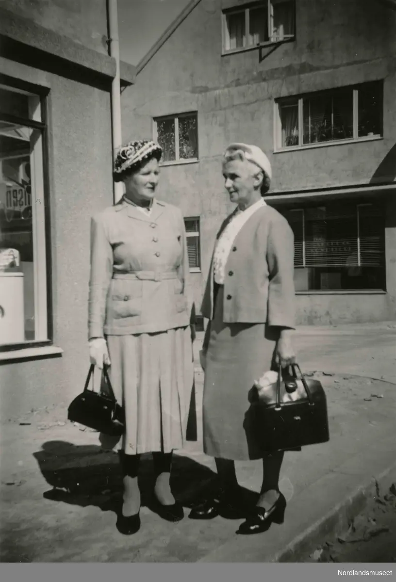 To eldre kvinner med todelte drakter, hatter og håndvesker. De står ute på en gate. Klara Simonsen, Bodø, til høyre.