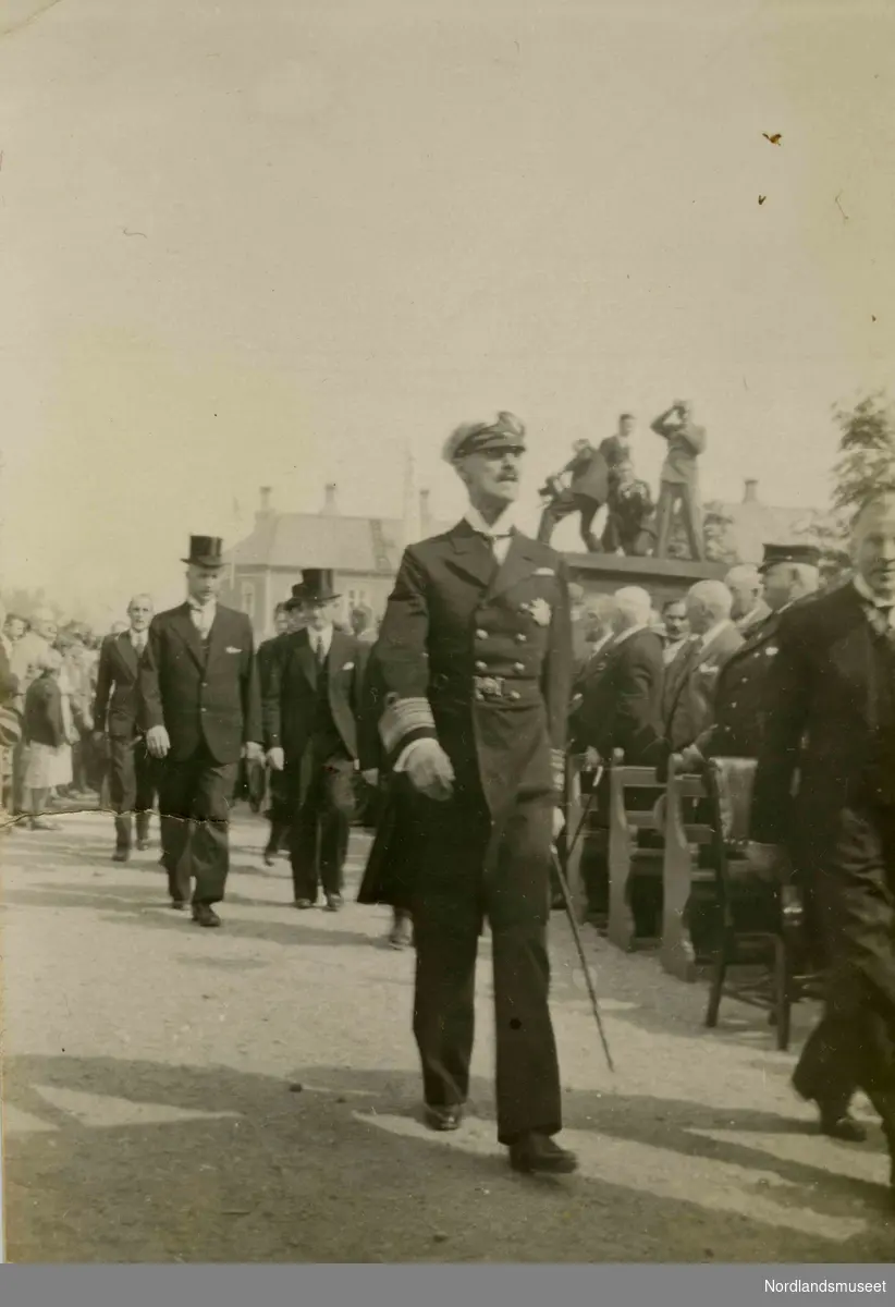Kong Haakon VII. I bakgrunnen ser vi flere menn iført dress og flosshatt. Benker, tilskuere og fotografer sees til høyre i bildet.