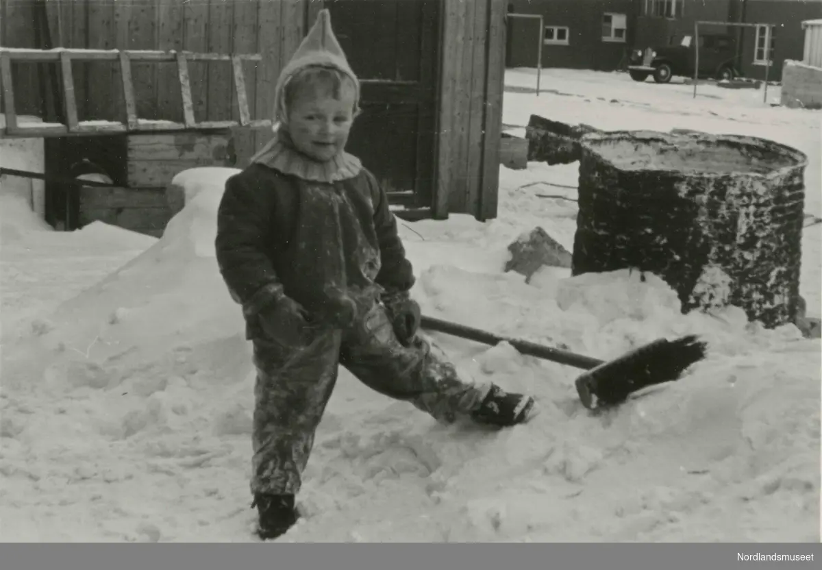 Et lite barn med strikket lue står ute i snøen. Bildetekst: Bakgård i Dronningens gate 3".