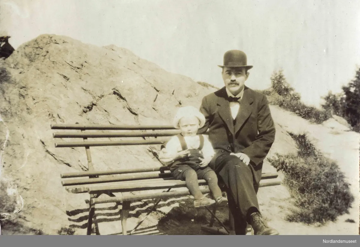 En mann og et lite barn sitter på en trebenk. Et berg i bakgrunnen. Mannen har på seg dress og bowlerhatt.