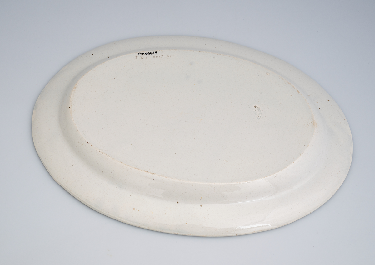 Et lite ovalt serveringsfat av glasert keramikk/porselen. Det er ensfarget hvitt (med hint av lyst blågrått i) på farge. Det er et enkelt fat uten særlig dekor. Det er stemplet på undersiden.