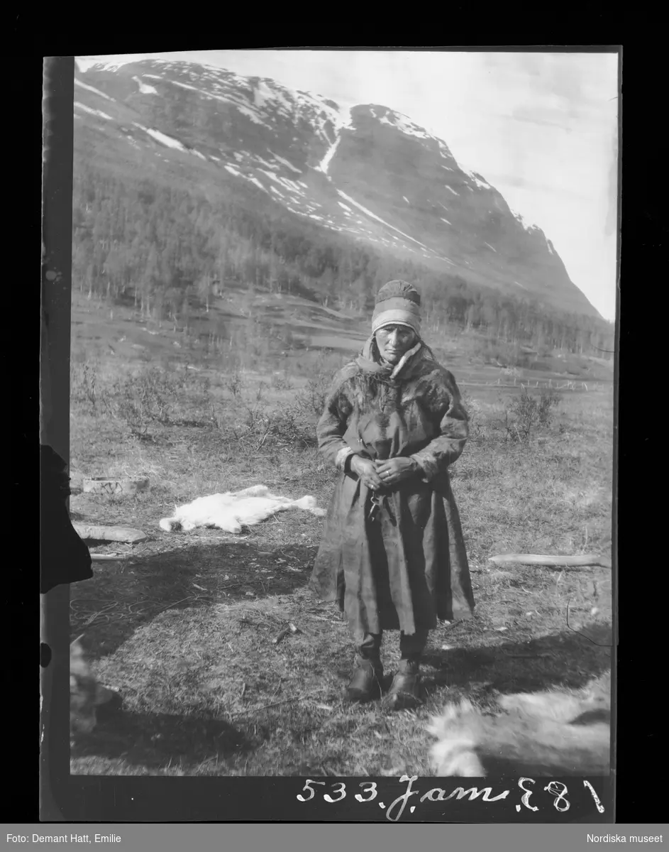 Kvinna, Girste, enligt bildtext gift med Andaras, står i landskap med fjäll i bakgrunden. På marken ligger renskinn. Bilden ingår i en serie fotografier tagna av Emilie Demant Hatt i Sapmi mellan åren 1907 och 1916.