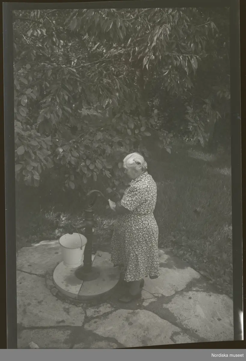 En kvinna hämtar vatten i en spann från en pump på gården.