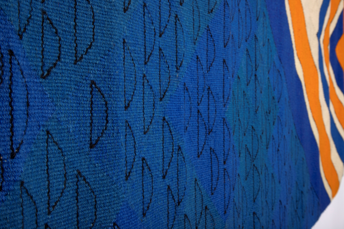 Håndvevd rektangulært billedvev i ull med geometrisk motiv i svart, blått, oransje og beige. Langs venstre og høyre kant er renningstrådene flettet og festet på baksiden med påsydde vevde bånd.