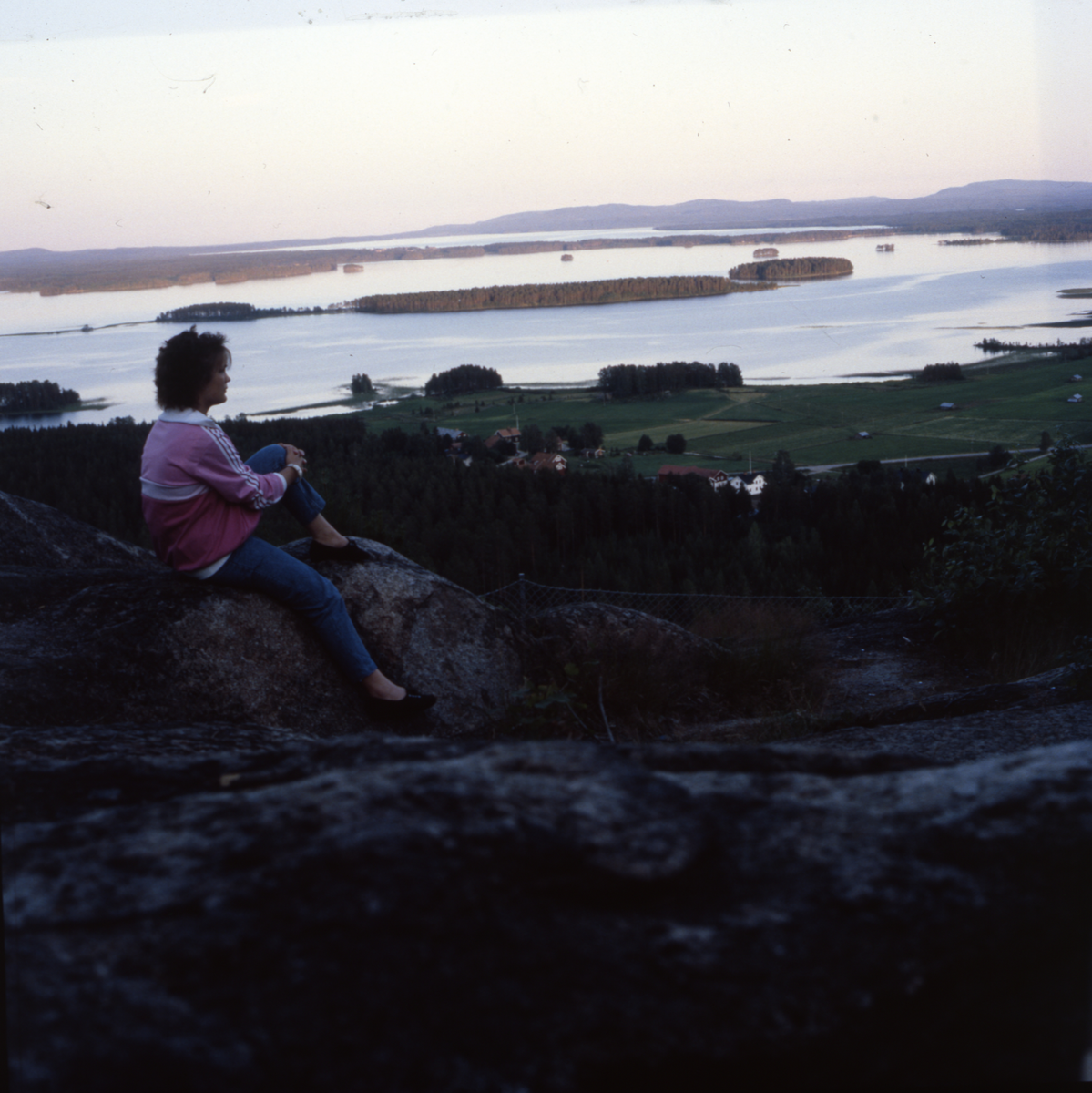 En kvinna sitter på en sten och blickar ut över ett landskap med åkrar, byggnader, sjö och berg.