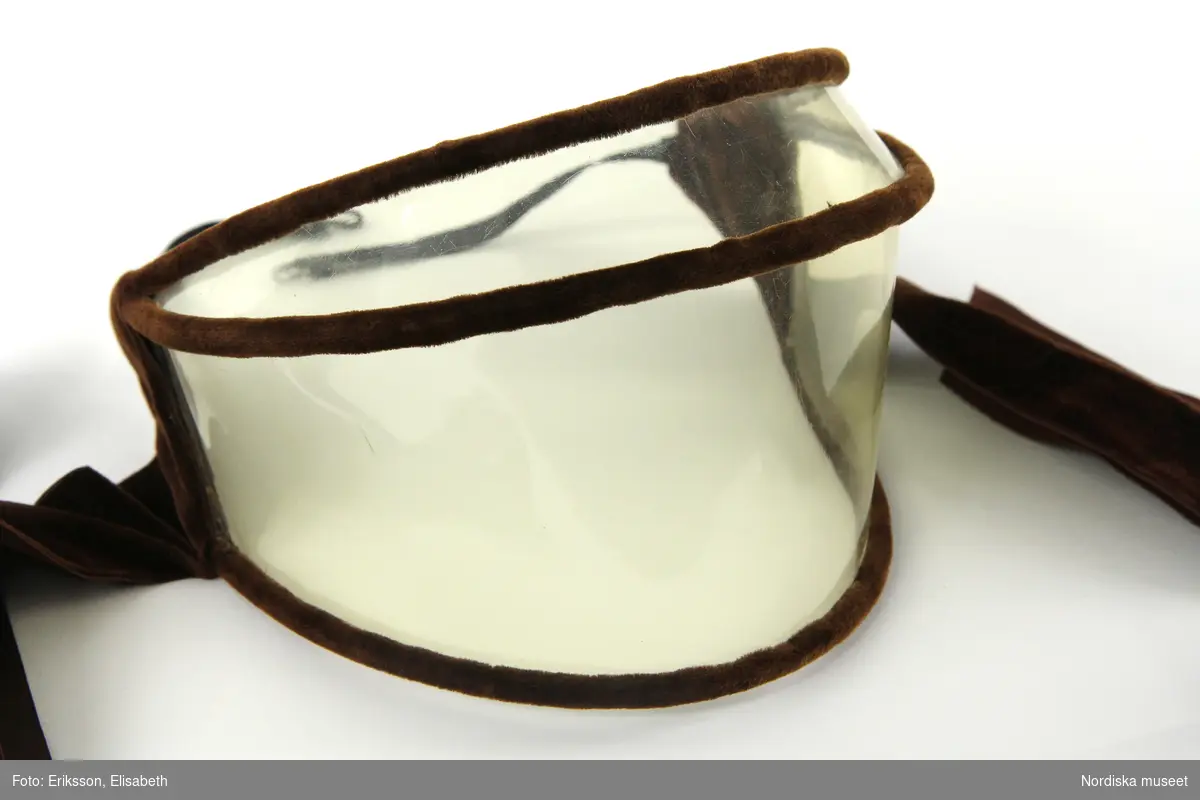 Bilglasögon i form av en skärm av transparent celluloid delad i två delar. Kantad med brun sammet. Baktill bruna, breda och långa sammetsband för ihopfästning under hakan, samt smala svarta resårband med hake och hyska för ihopfästning i nacken. 
/Leif Wallin 2024-04-04